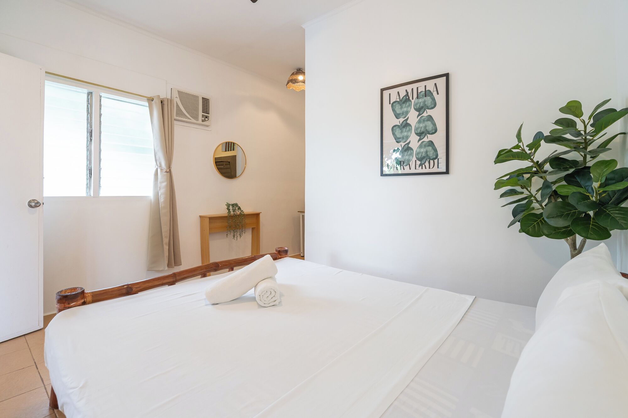 Una cama bien hecha con una toalla enrollada, una mesita de noche con un espejo, una obra de arte enmarcada en la pared y una planta en maceta.