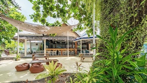 An outdoor seating area with wooden tables and benches, surrounded by lush greenery and a thatched roof structure.