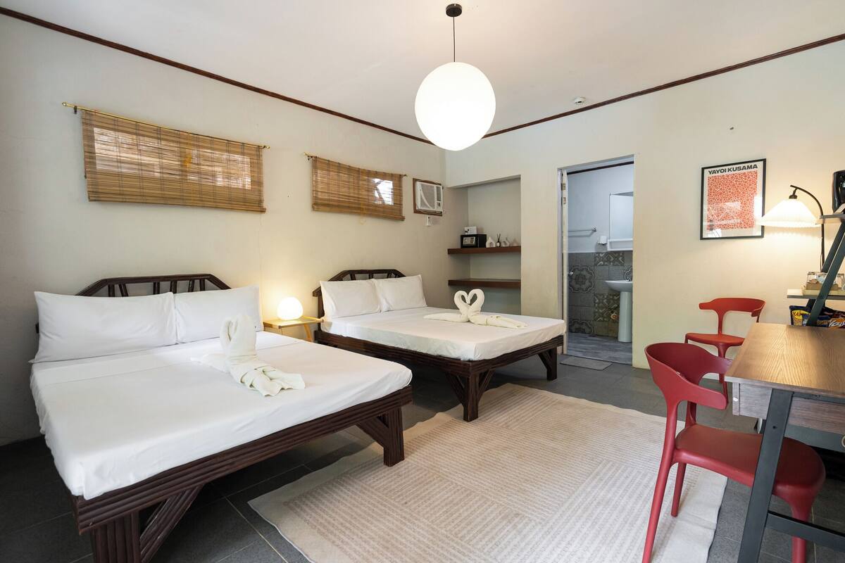 Une chambre d’hôtel avec deux lits, une table à manger avec des chaises rouges, un luminaire suspendu et une salle de bain visible à travers une porte ouverte.