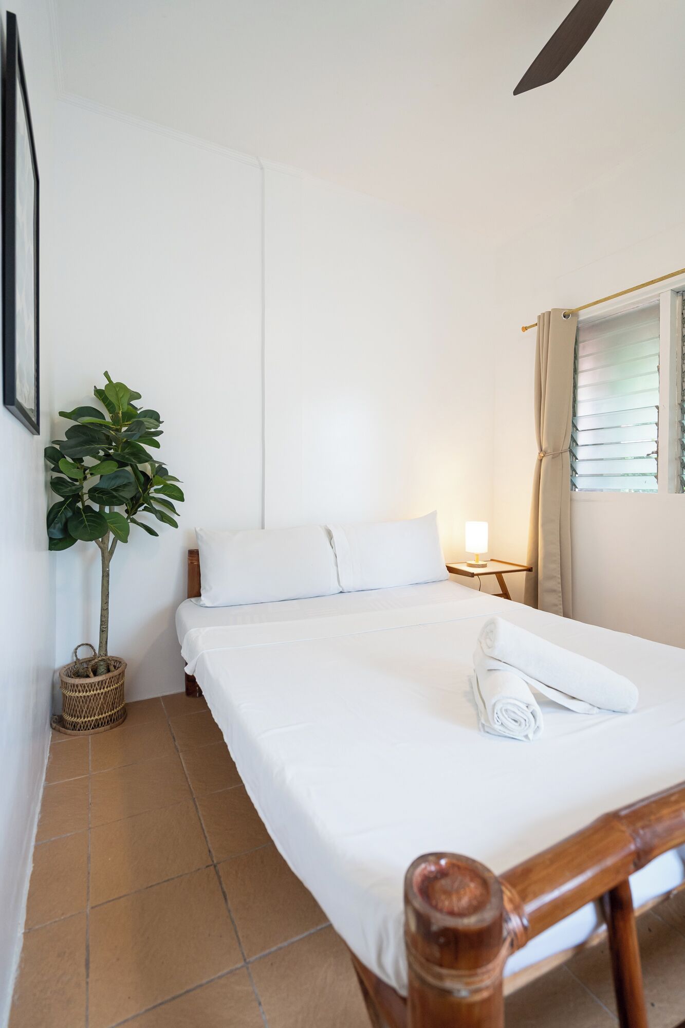 A small, neatly made bed with white linens, a bedside lamp, and a potted plant in a wicker basket.