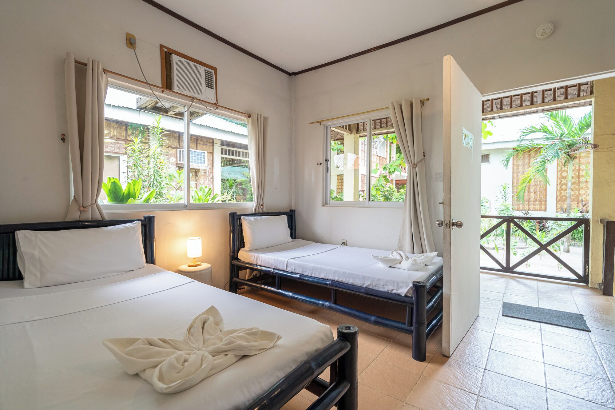 A room with two beds, a window with curtains, a bedside lamp, and a view of the outside.