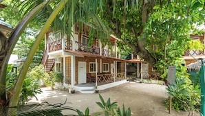 A two-story building with a balcony, surrounded by lush greenery and a sandy ground.