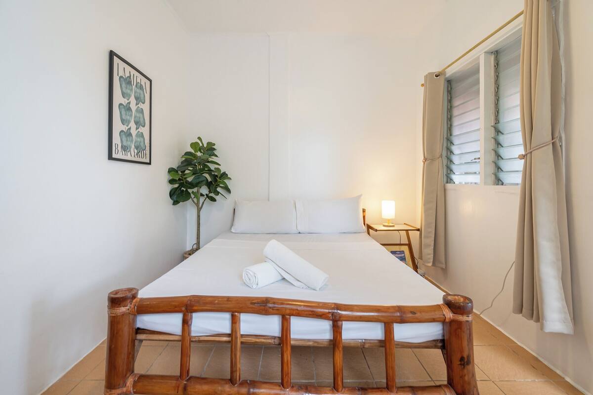 Un lit bien fait, avec du linge de lit blanc, une tête de lit en bois, une lampe de chevet, un tableau encadré au mur et une plante en pot.