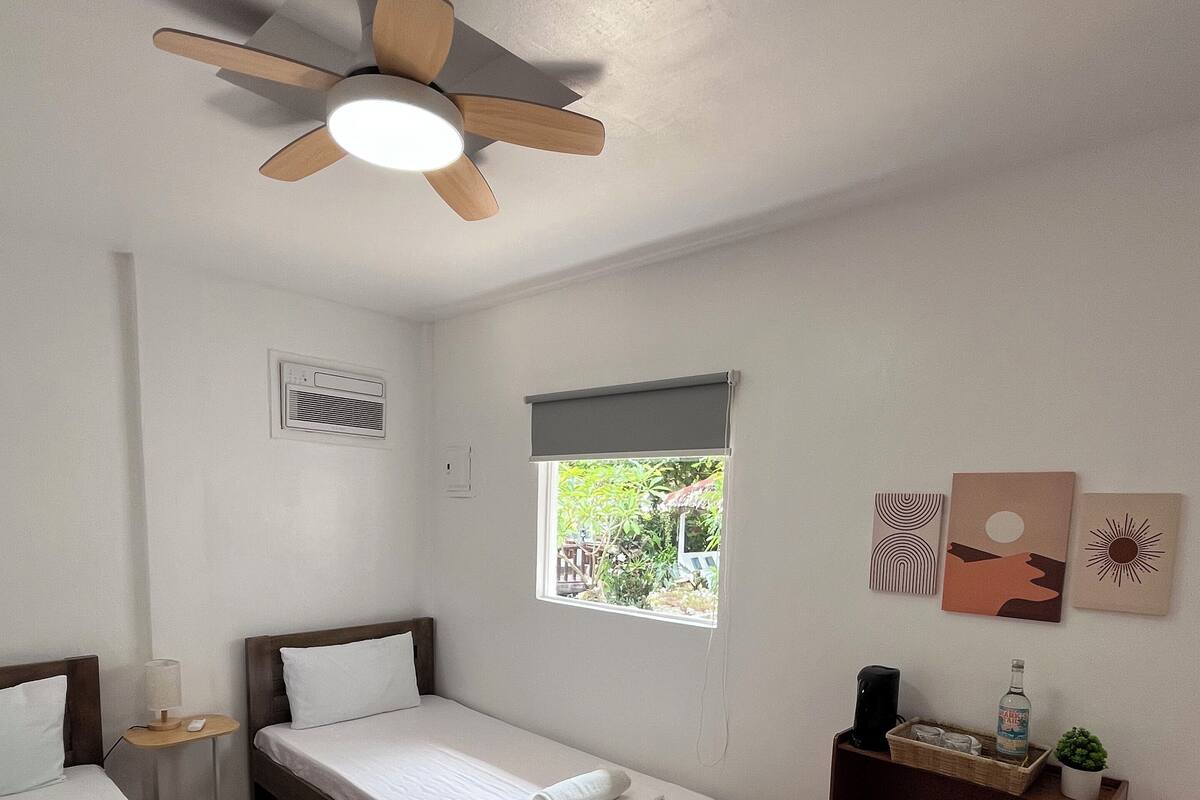 Une chambre avec deux lits simples, un ventilateur de plafond, une fenêtre avec vue, une petite étagère avec quelques objets et un climatiseur fixé au mur.