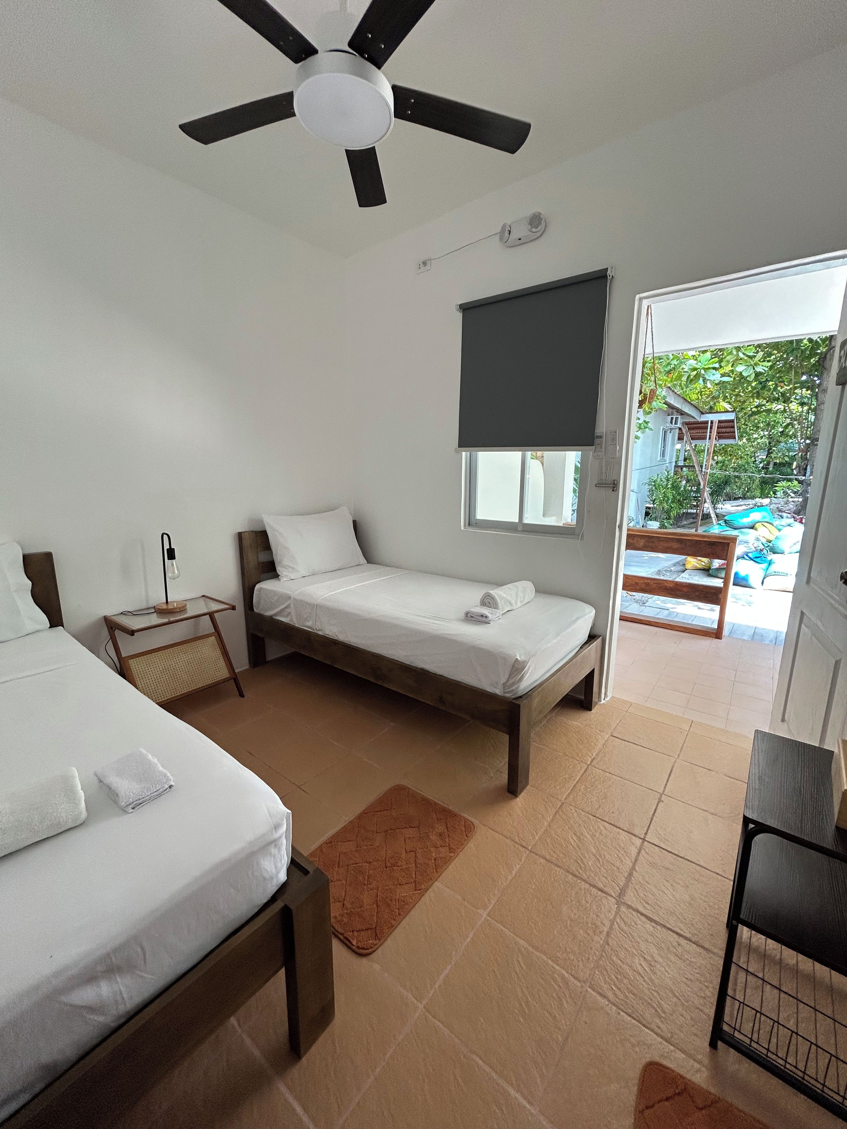 Un dormitorio con dos camas, un ventilador de techo, una ventana con persiana y vistas a un espacio exterior.