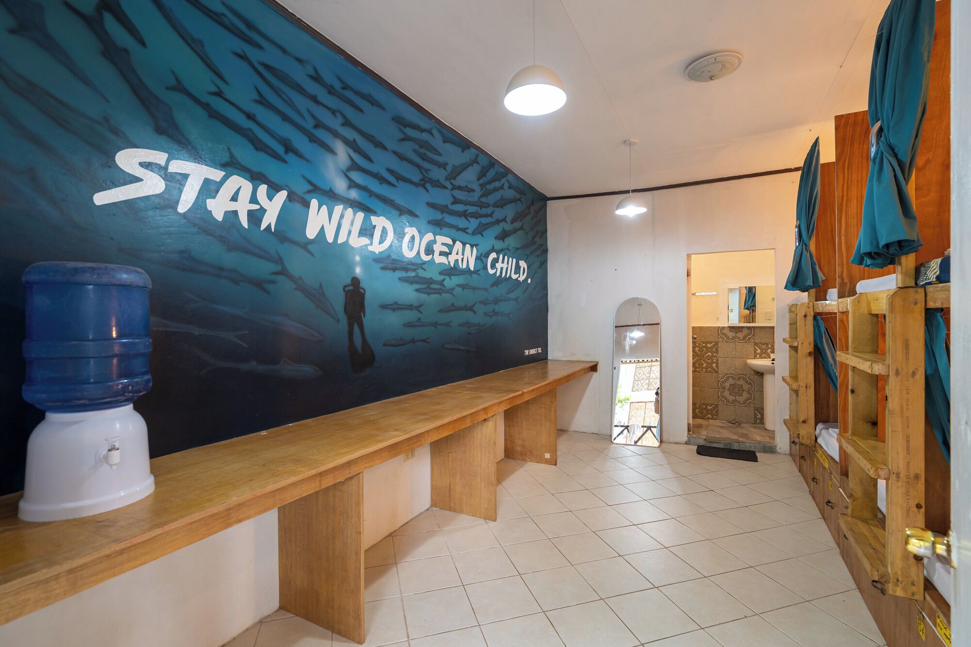 A bathroom with a mural of a diver and sharks, a wooden bench, a water dispenser, and wooden lockers.