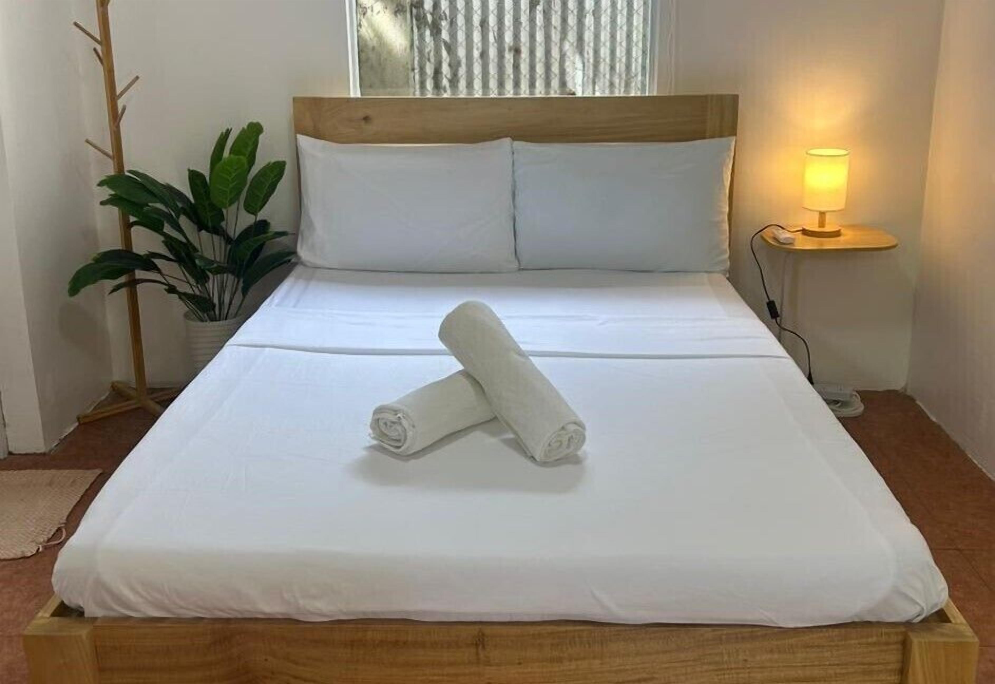Un lit bien fait, avec du linge de lit blanc et deux serviettes enroulées, une lampe de chevet et une plante en pot dans un coin.