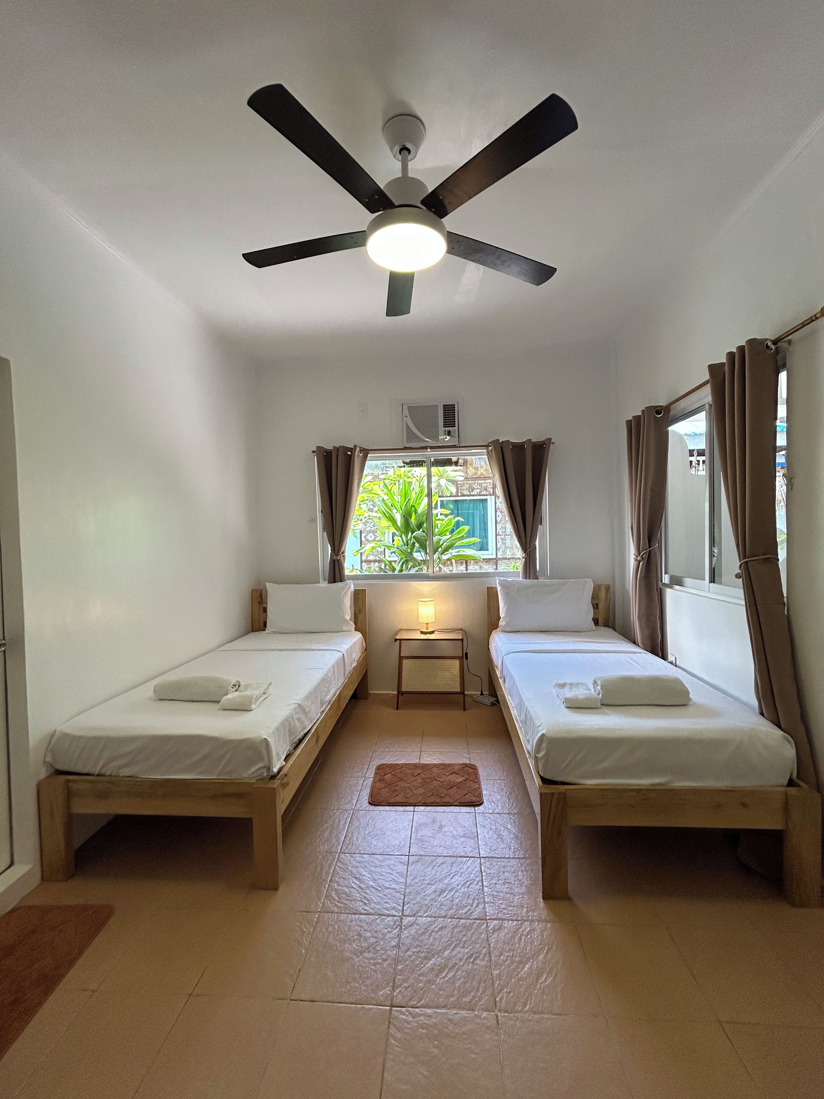 A small, single-room accommodation with two single beds, a ceiling fan, a window with curtains, and a small table with a lamp.