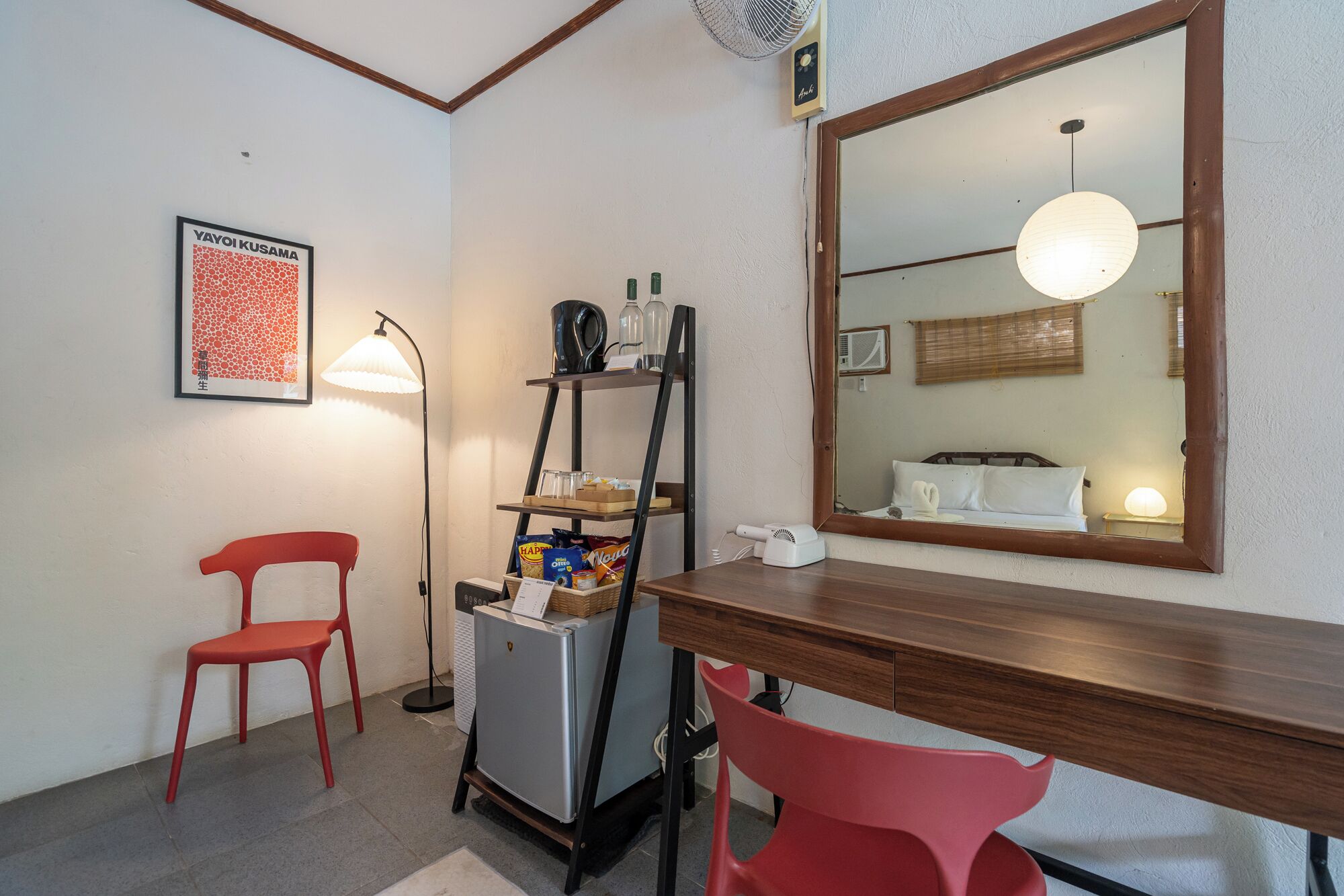 A compact room with a wooden desk, a red chair, a small refrigerator, a shelf with snacks, a lamp, and a large mirror reflecting the room.