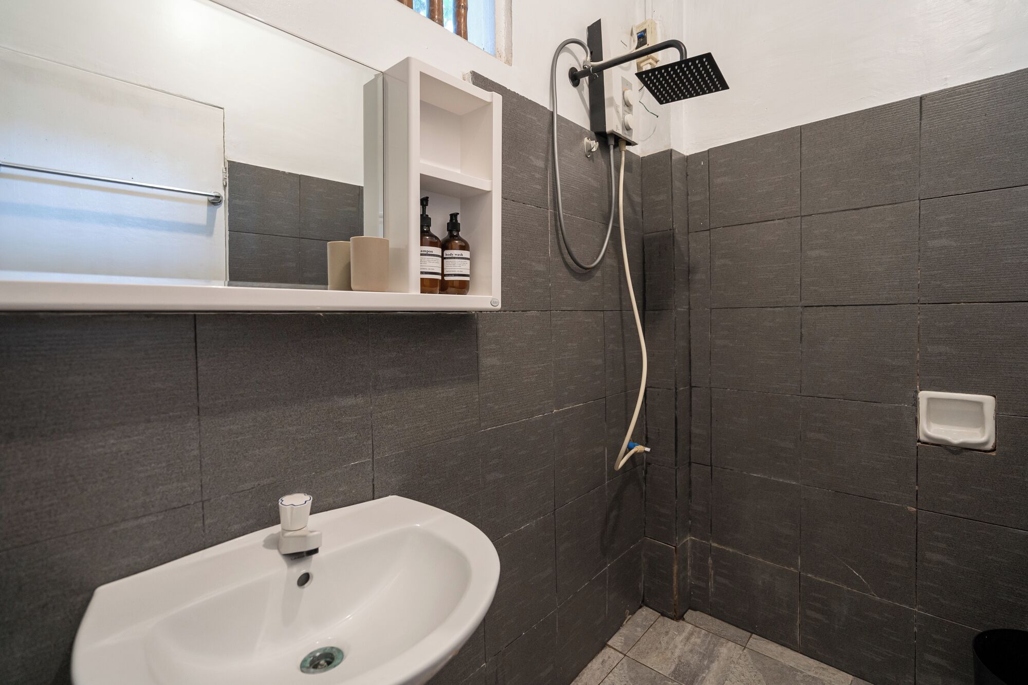 Een badkamer met een wit wastafel, donkere betegelde wanden, een douche met een handdouche en een plank met toiletartikelen.