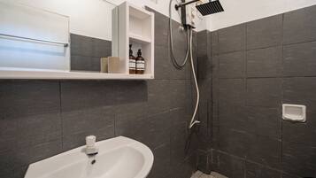 Un baño con un lavabo blanco, paredes de azulejos oscuros, ducha con cabezal de ducha manual y estante con artículos de aseo.