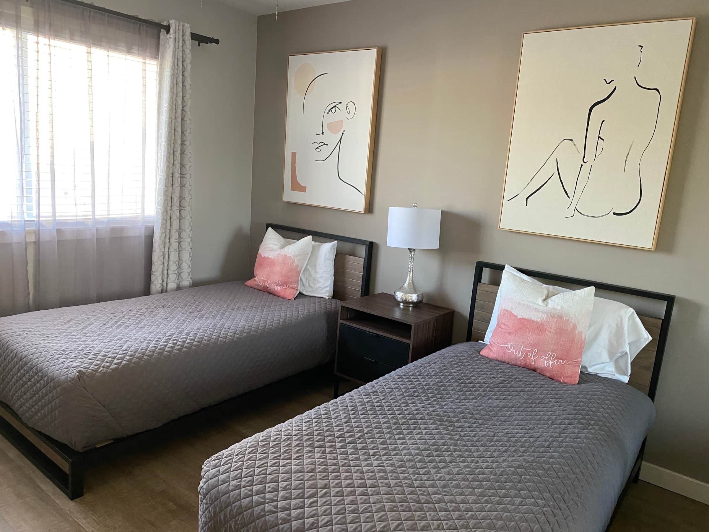 A hotel room with two single beds, each with a pillow and a bedside table with a lamp. There are abstract paintings on the walls and a window with sheer curtains.