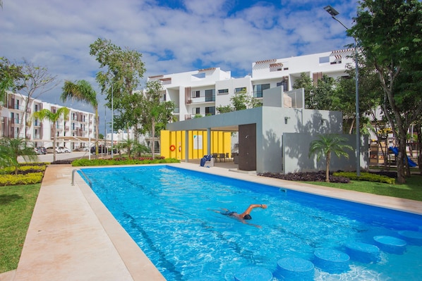 A swimming pool with a person swimming, surrounded by greenery and modern buildings.