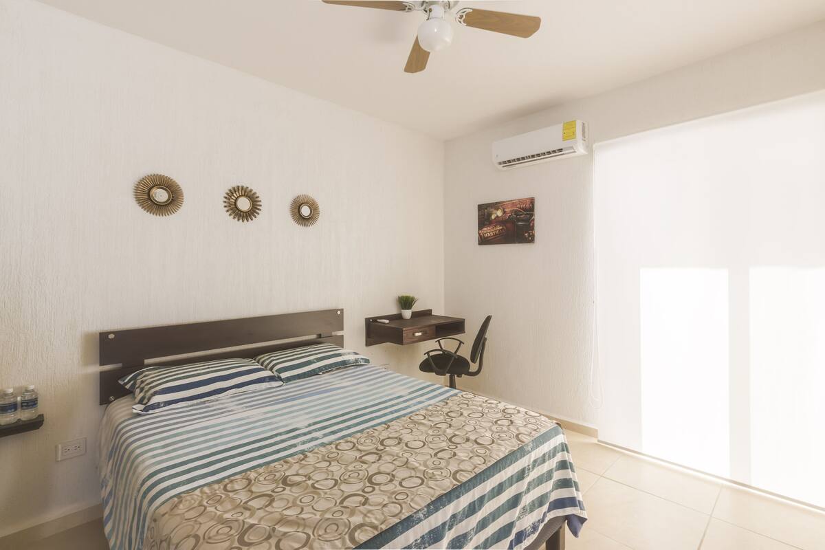 Un dormitorio con una cama, un ventilador de techo, un estante en la pared con una planta y una ventana.