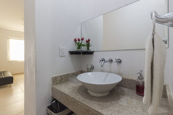 Casa de banho com lavatório branco, espelho, porta-toalhas e uma pequena estante com flores vermelhas.