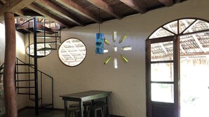 Ein Zimmer mit Wendeltreppe, einem Rundfenster, einem Tisch mit Hocker und einer sichtbaren Tür mit Strohdach durch das Fenster hindurch.