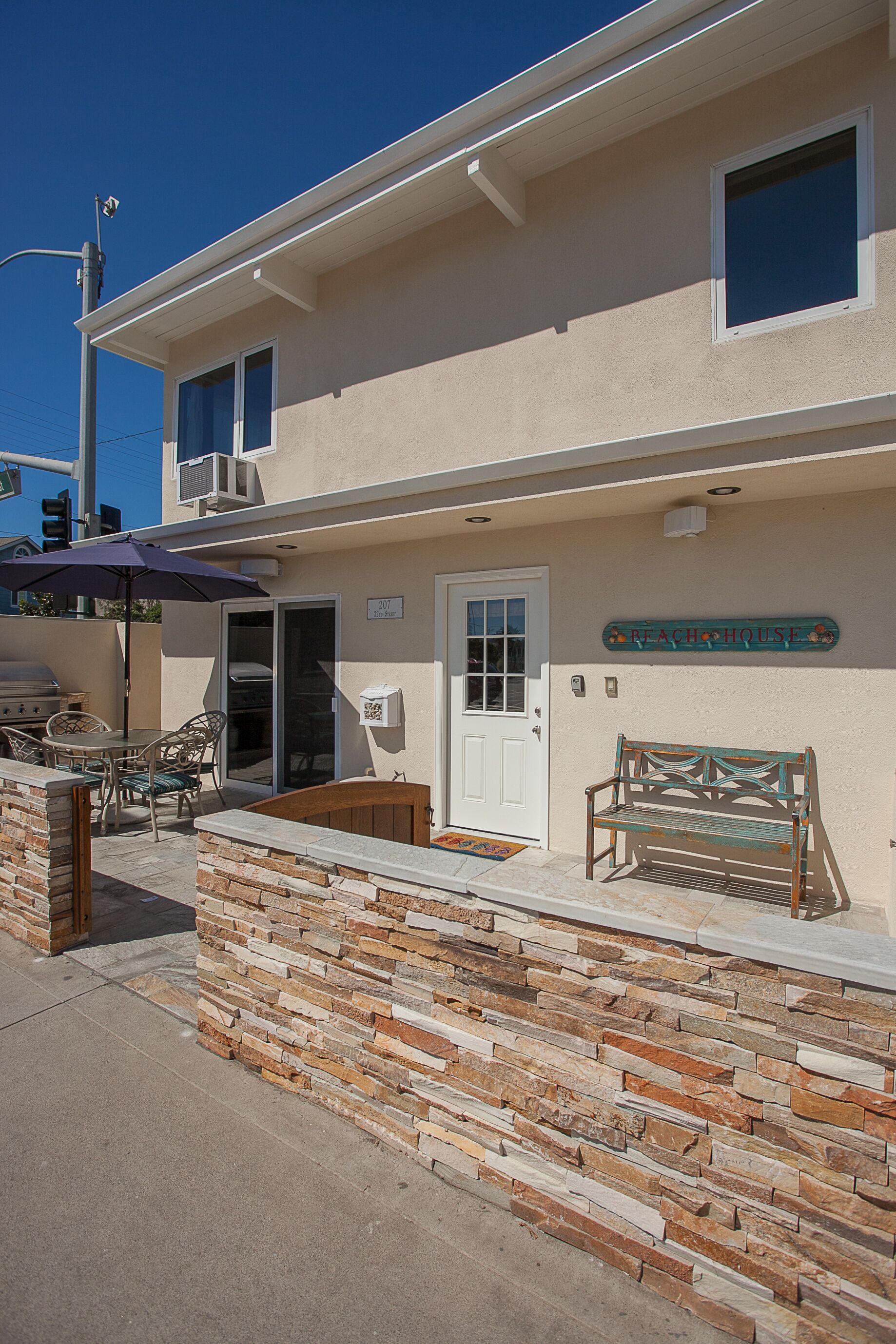A building with a stone wall, a wooden bench, a table with chairs, and a sign that reads "Beach House".
