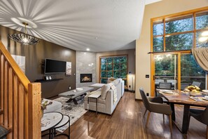 A modern living room with a fireplace, a dining area, and large windows offering a view of trees.