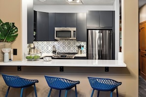A modern kitchen with a microwave, refrigerator, and patterned backsplash. There are three blue chairs in front of the counter.