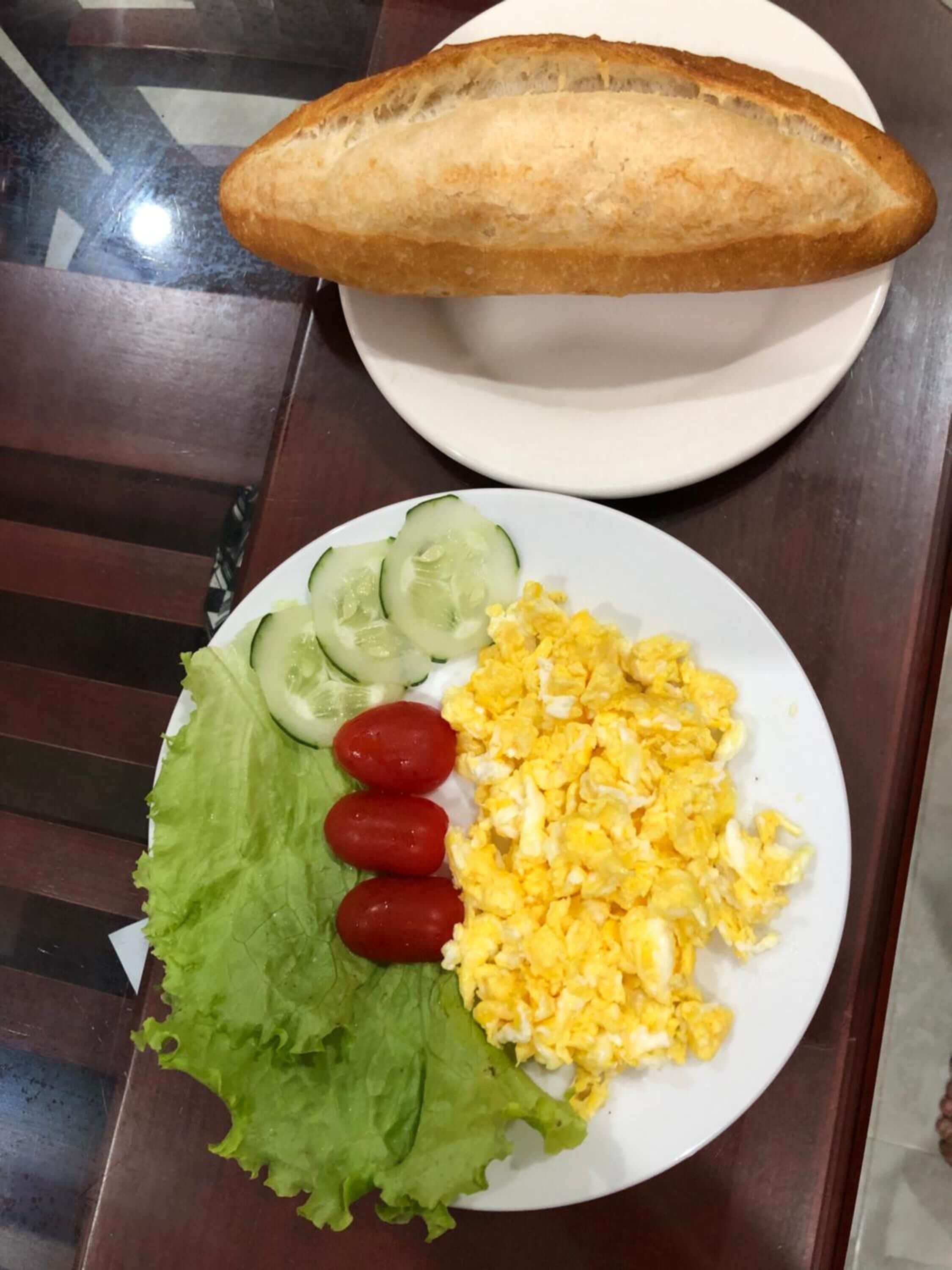A plate with scrambled eggs, sliced cucumbers, lettuce, and cherry tomatoes, accompanied by a baguette on a white plate.