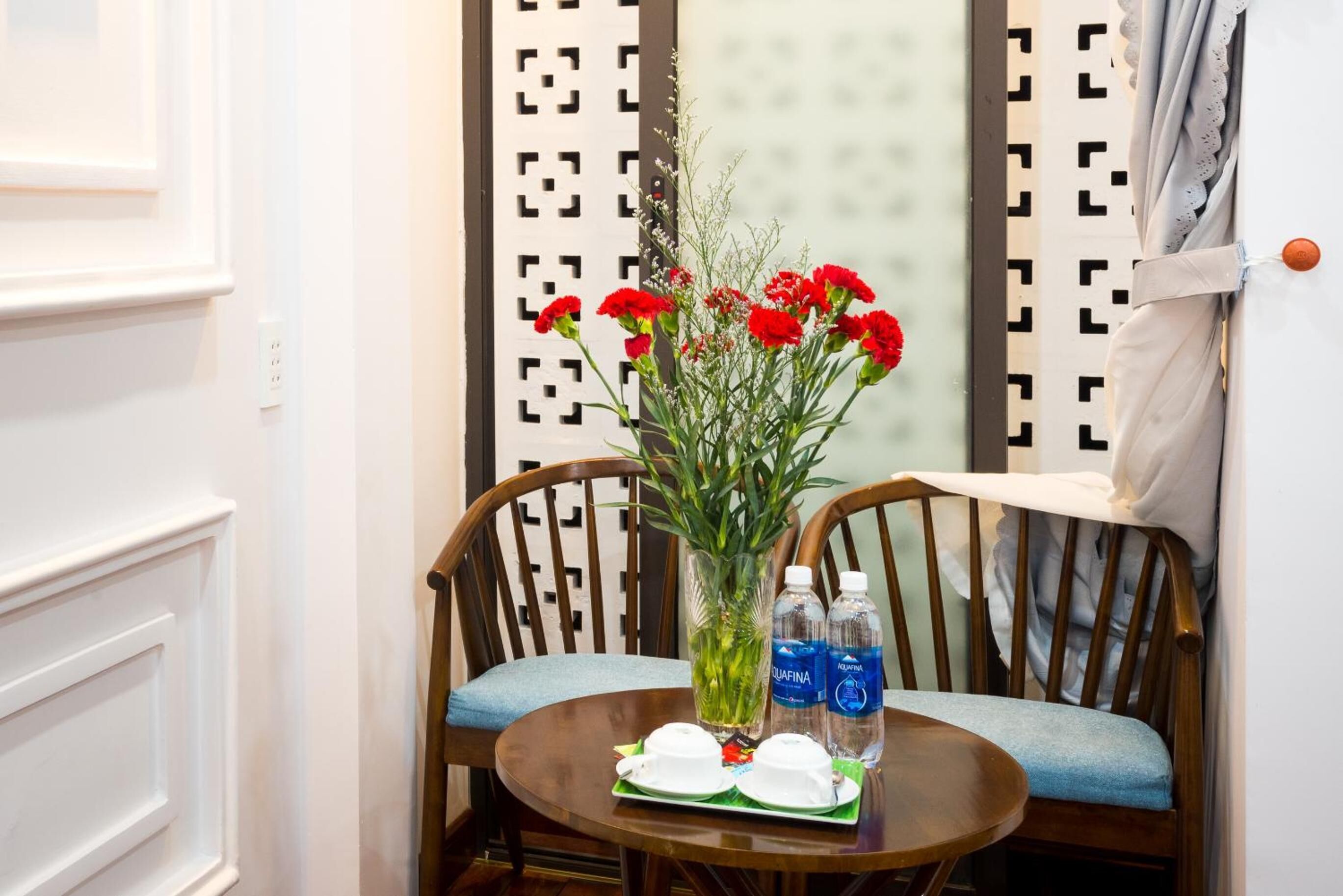 A corner of a room with a wooden chair, a small round table with a vase of red flowers, two water bottles, and a green tray.