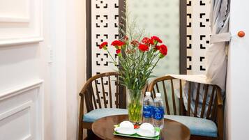 Un coin de pièce avec une chaise en bois, une petite table ronde sur laquelle se trouve un vase de fleurs rouges, deux bouteilles d'eau et un plateau vert.