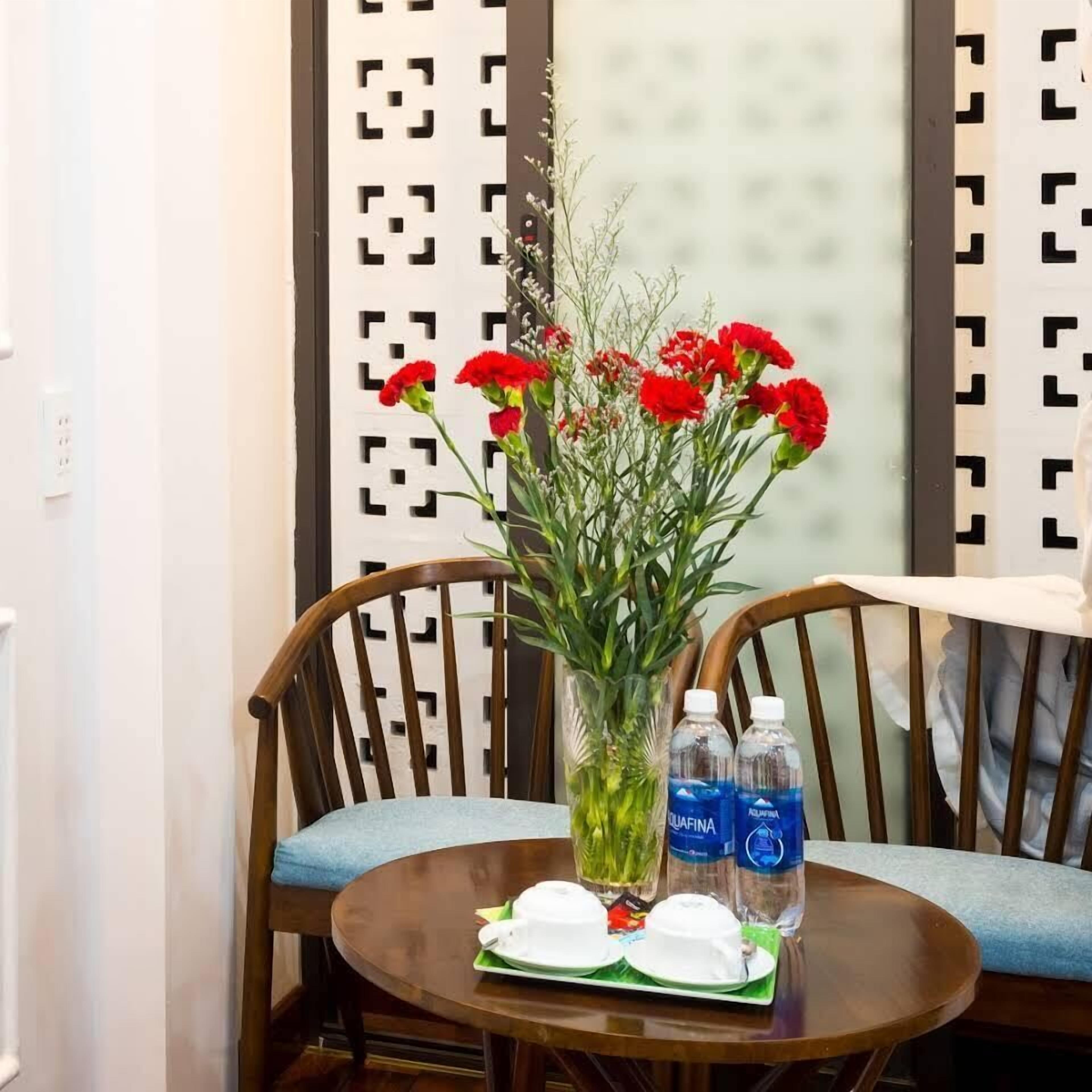 Una habitación con una silla de madera, una mesita pequeña, un jarrón con flores rojas y dos botellas de agua.