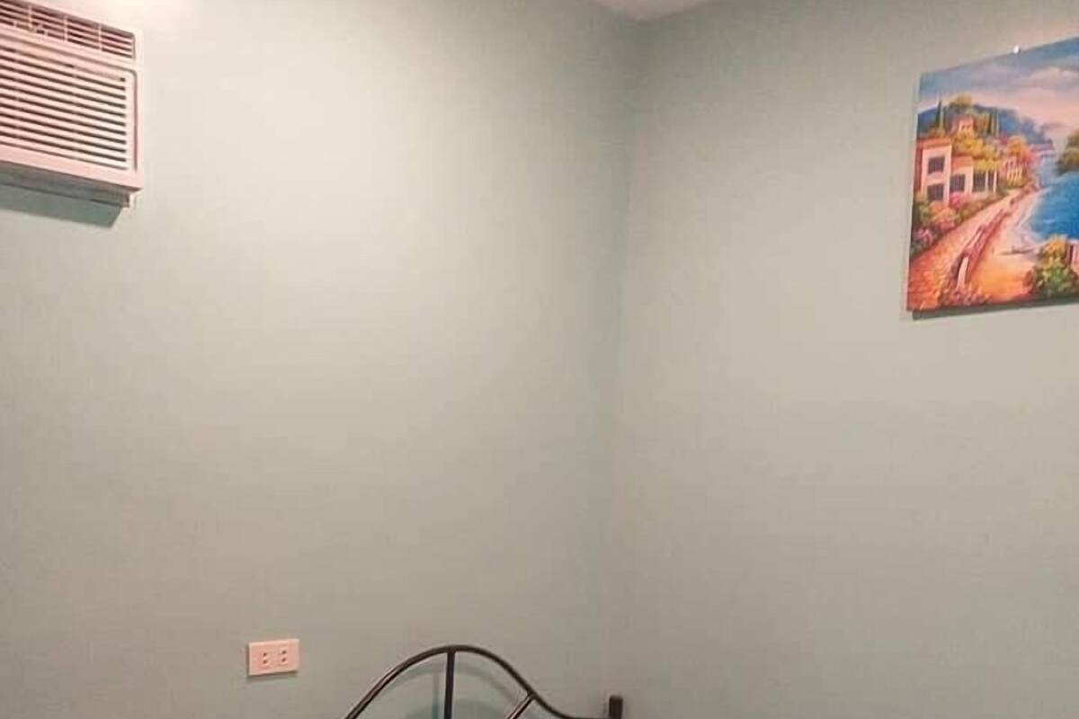 A small, single-bed room with a metal frame bed, a blue mattress, a small table, and a wall-mounted air conditioner.