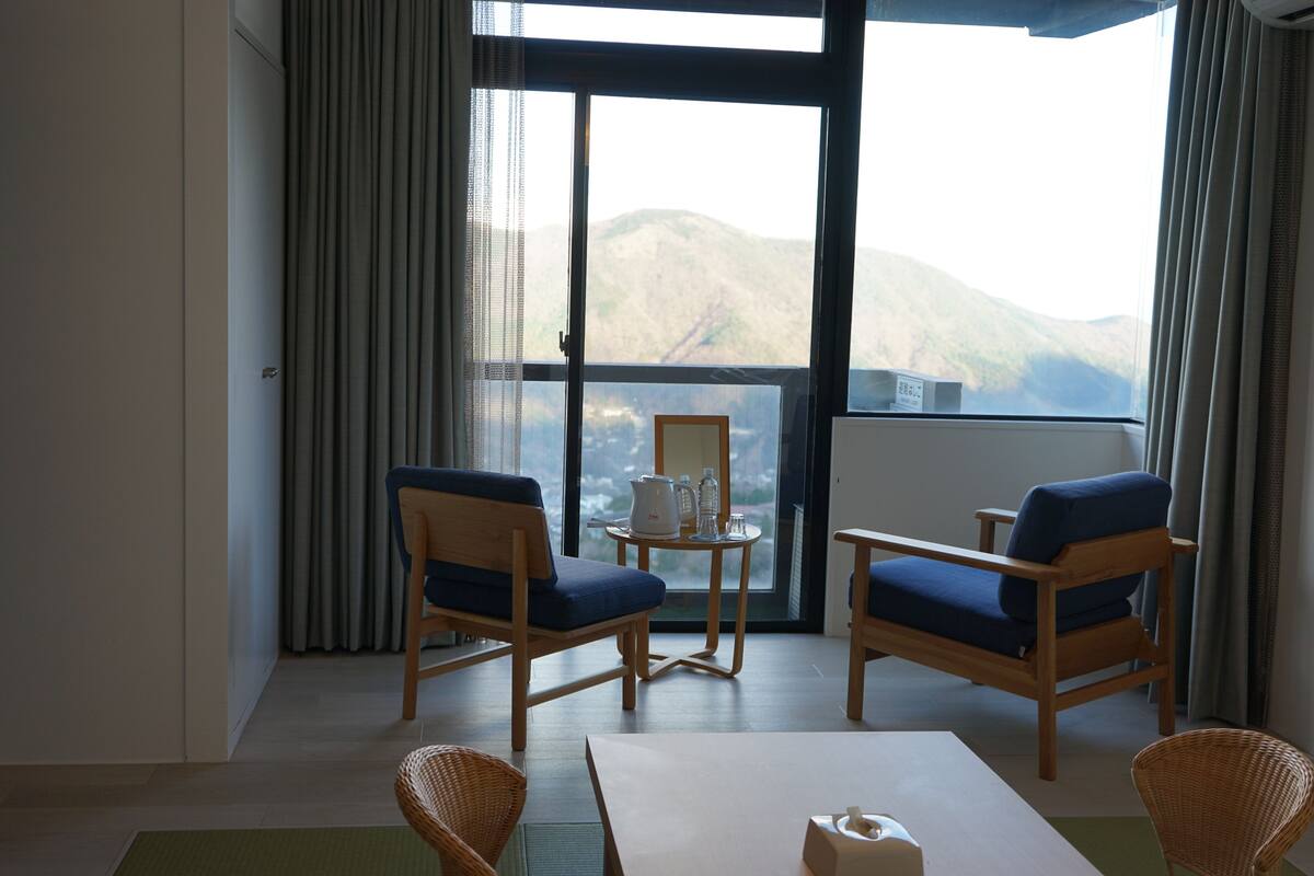 A room with two blue upholstered chairs, a small round table with a kettle and glasses, and a view of mountains through a large window.