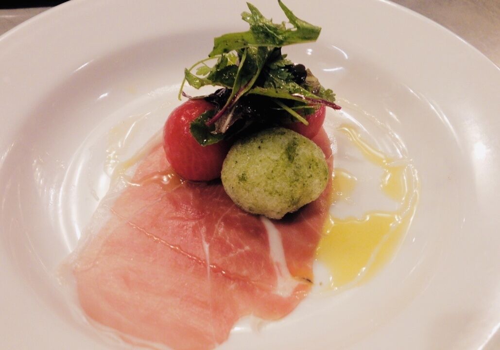 Un piatto con un pomodoro rosso, pesto verde e una fetta di carne rosa, forse prosciutto, su un piatto bianco.