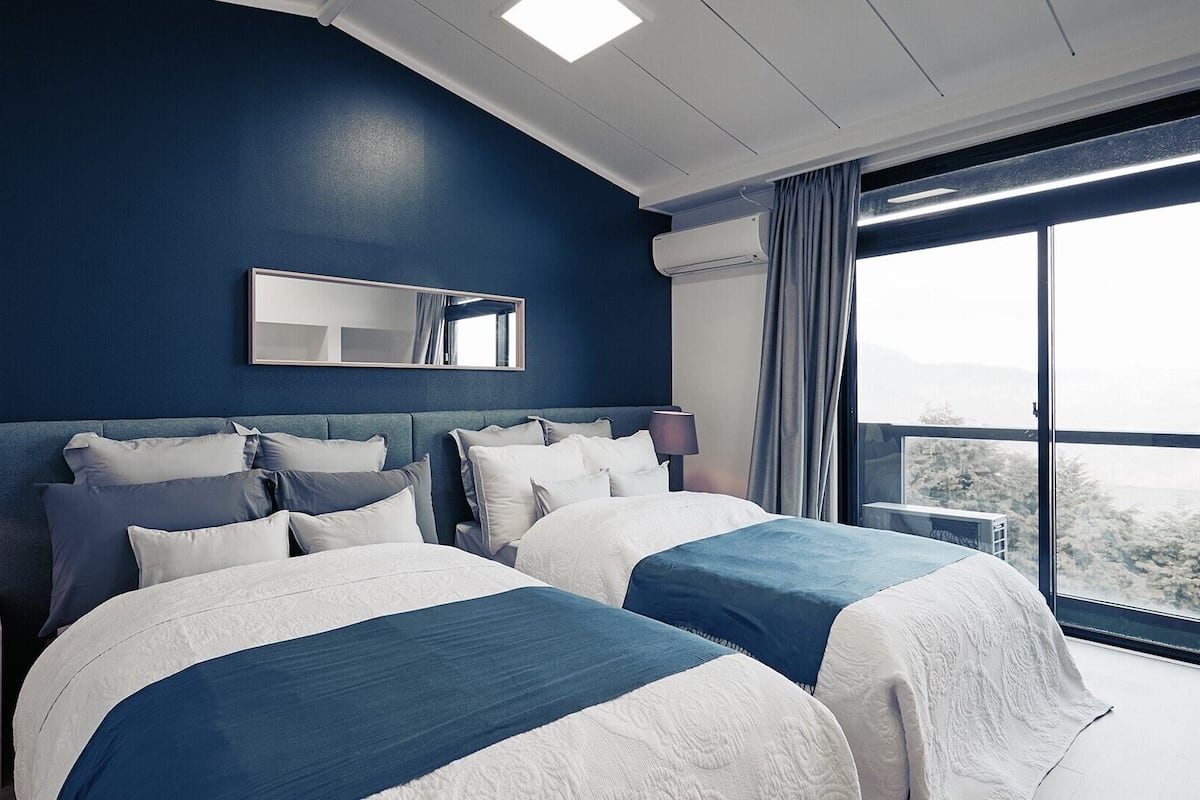 A modern hotel room with two beds, a large window with a view, a mirror on the wall, and an air conditioning unit.