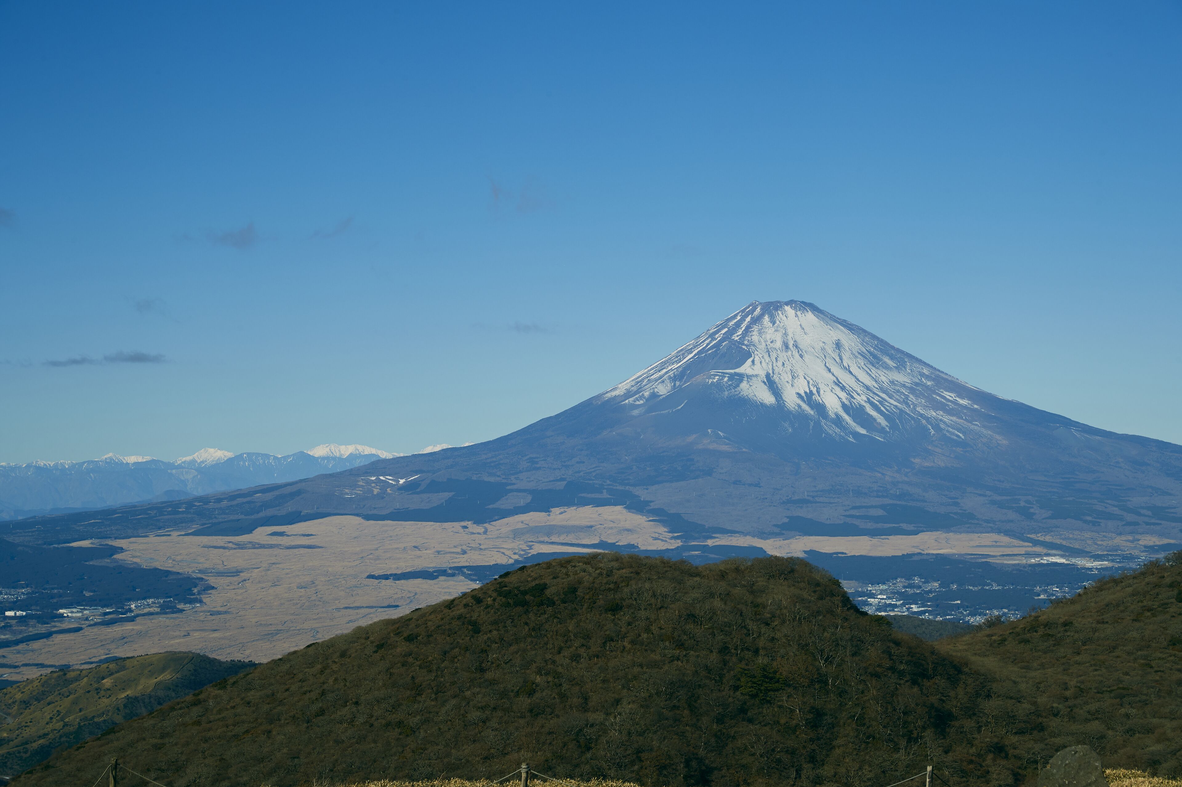 A mountain with a snow-capped peak, surrounded by forested hills and a valley with scattered buildings.