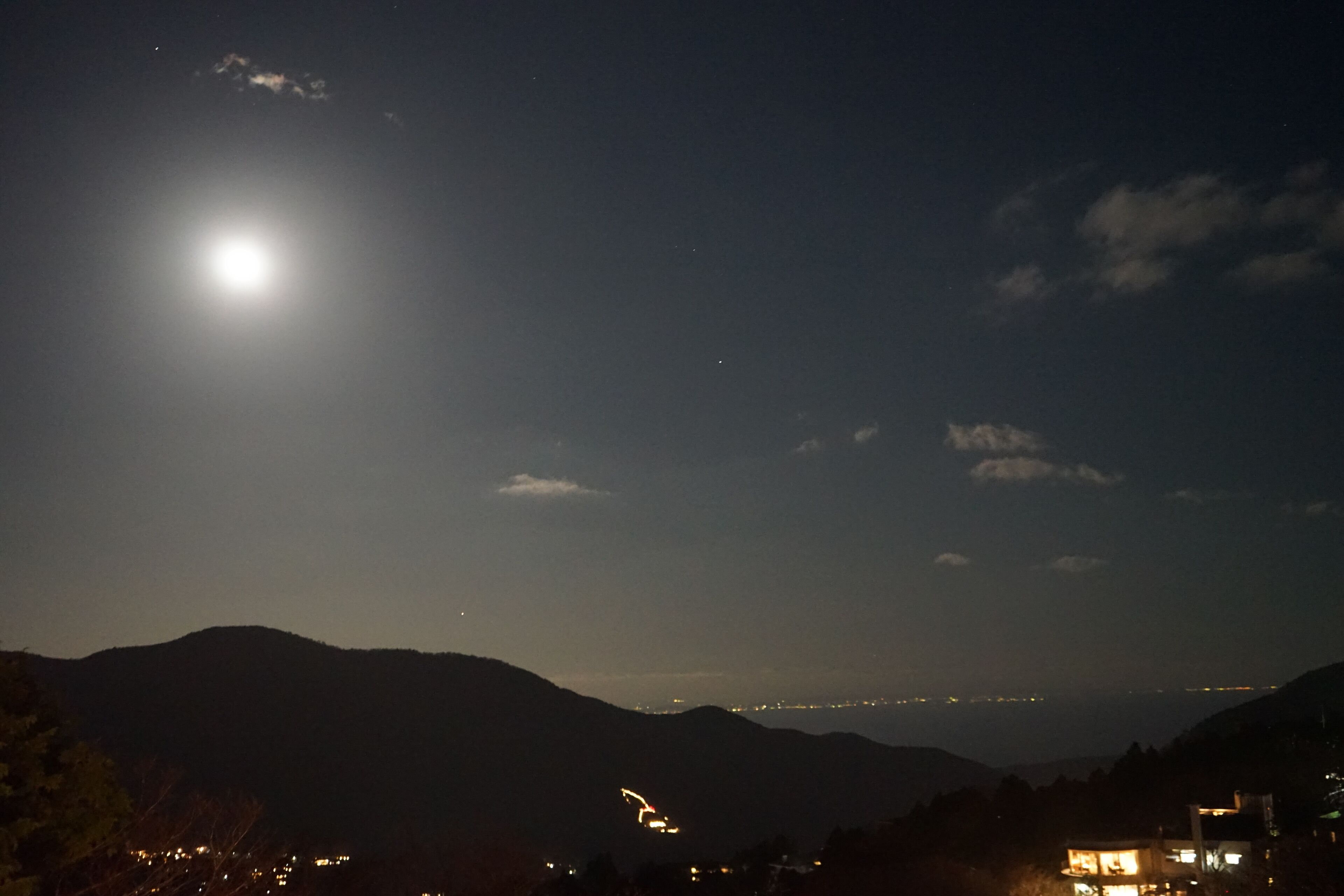 Vista nocturna de una zona montañosa con una luna brillante y luces de una ciudad a lo lejos.