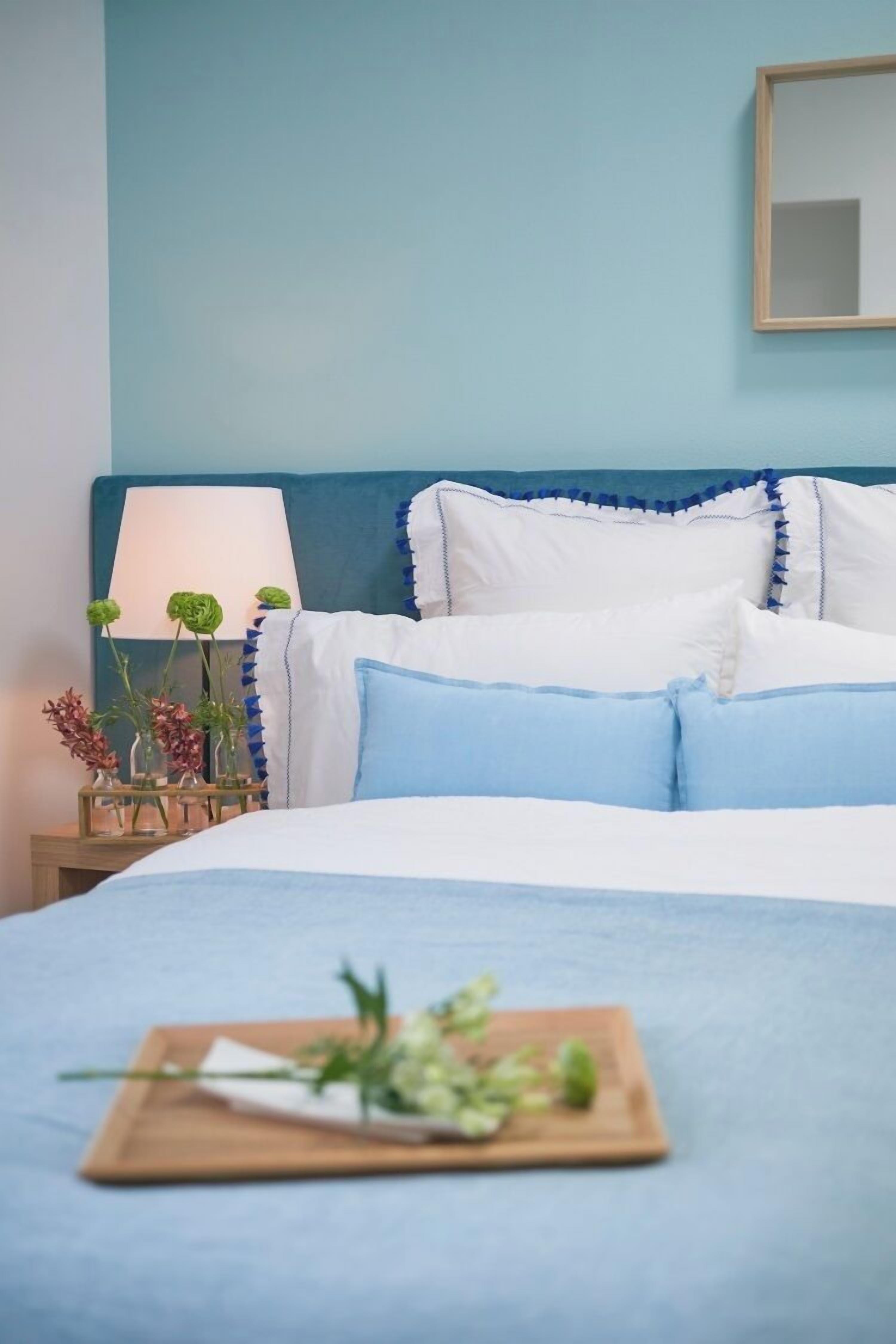 A neatly made bed with blue bedding, a bedside table with a lamp and decorative flowers, and a wooden tray with greenery.