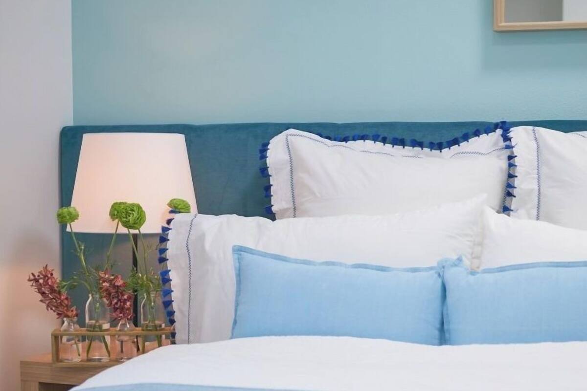 A neatly made bed with blue bedding, a bedside table with a lamp and decorative flowers, and a wooden tray with greenery.