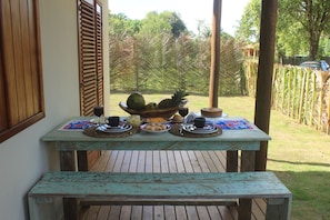Una mesa de madera rústica con un frutero, platos y tazas en un patio con vistas a un jardín y un coche al fondo.