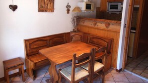 Ein Essbereich mit einer Holzbänke und einem Tisch, einem kleinen Hocker und einer Küchenzeile mit Holzschränken und Geräten.