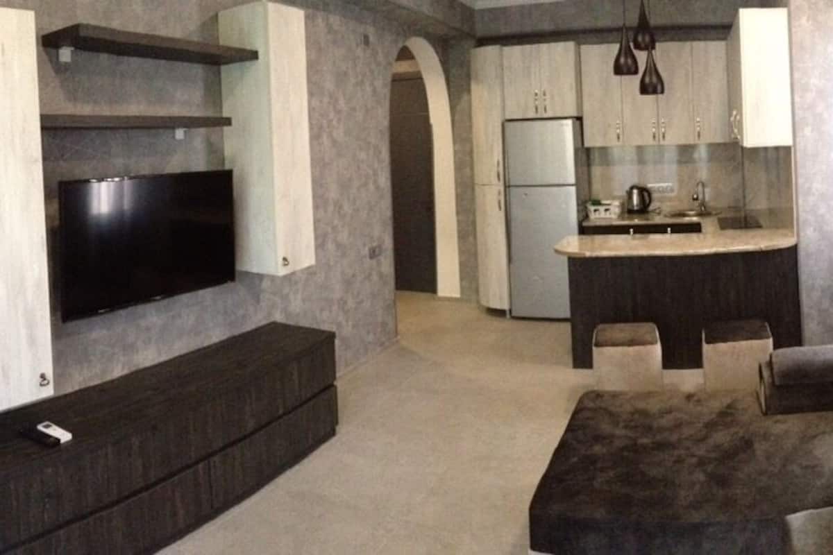 A modern living room with a flat-screen TV, built-in seating, and a kitchen area in the background.