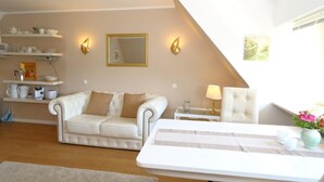 A living room with a white sofa, a small table, and a chair. There are wall-mounted shelves with kitchen items and a mirror. - Teewelkenhüs sun apartment (Sylt)