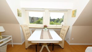 A dining area with a white table, two beige chairs, a vase with flowers, and a view of greenery through large windows. - Teewelkenhüs sun apartment (Sylt)