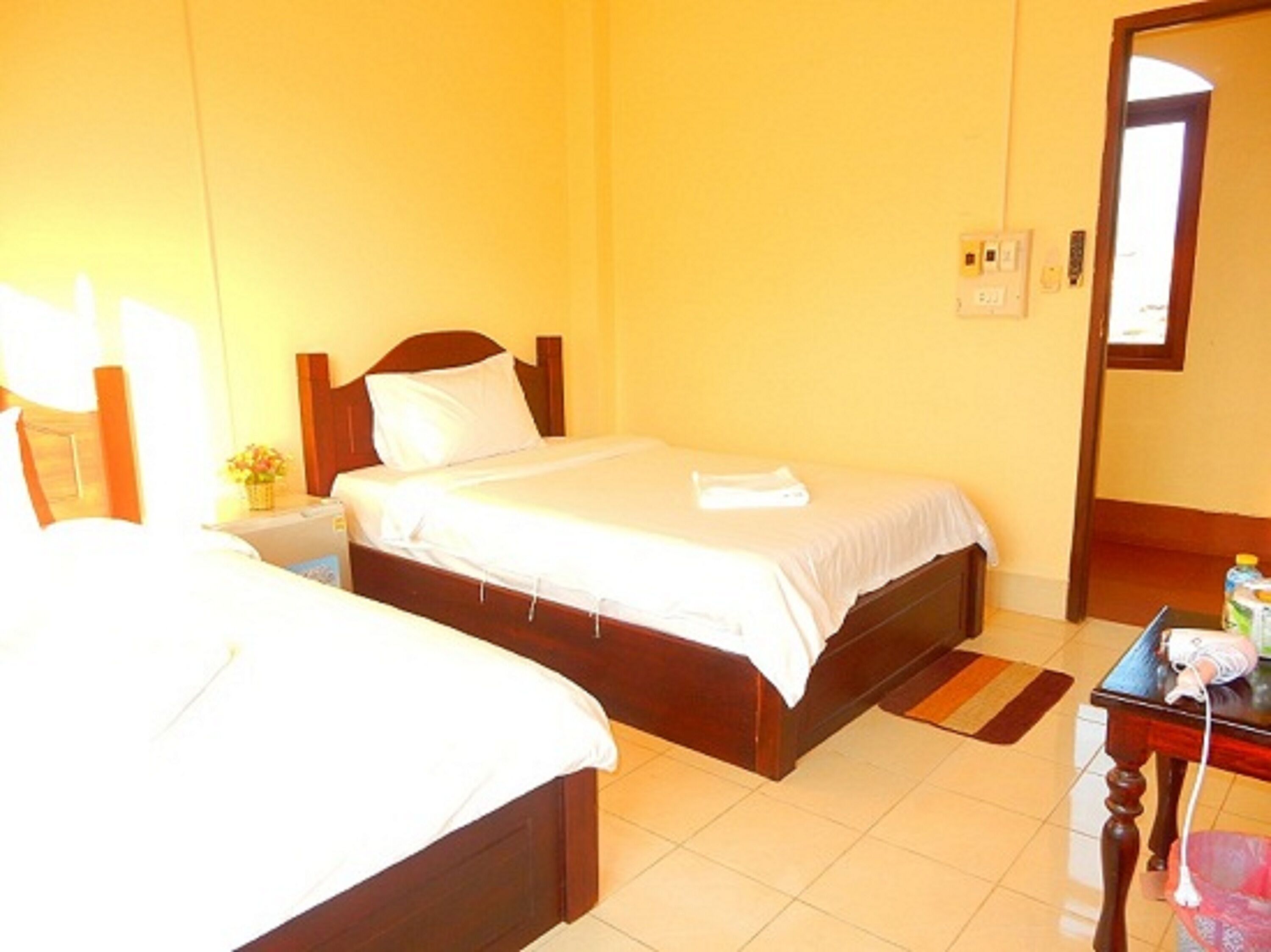 A hotel room with two single beds, a bedside table with a lamp, a small table with a vase of flowers, and a door leading to another room.
