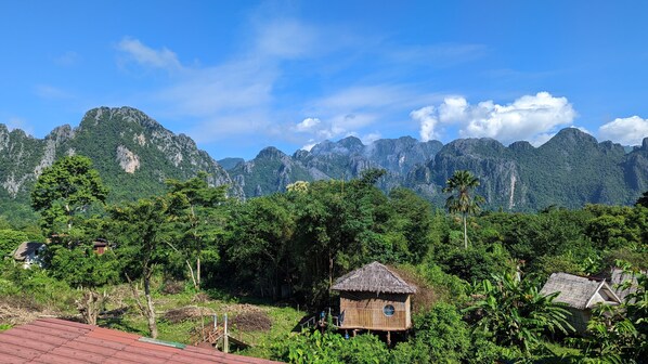 A rural landscape with a thatched-roof hut surrounded by lush greenery and mountains in the background.