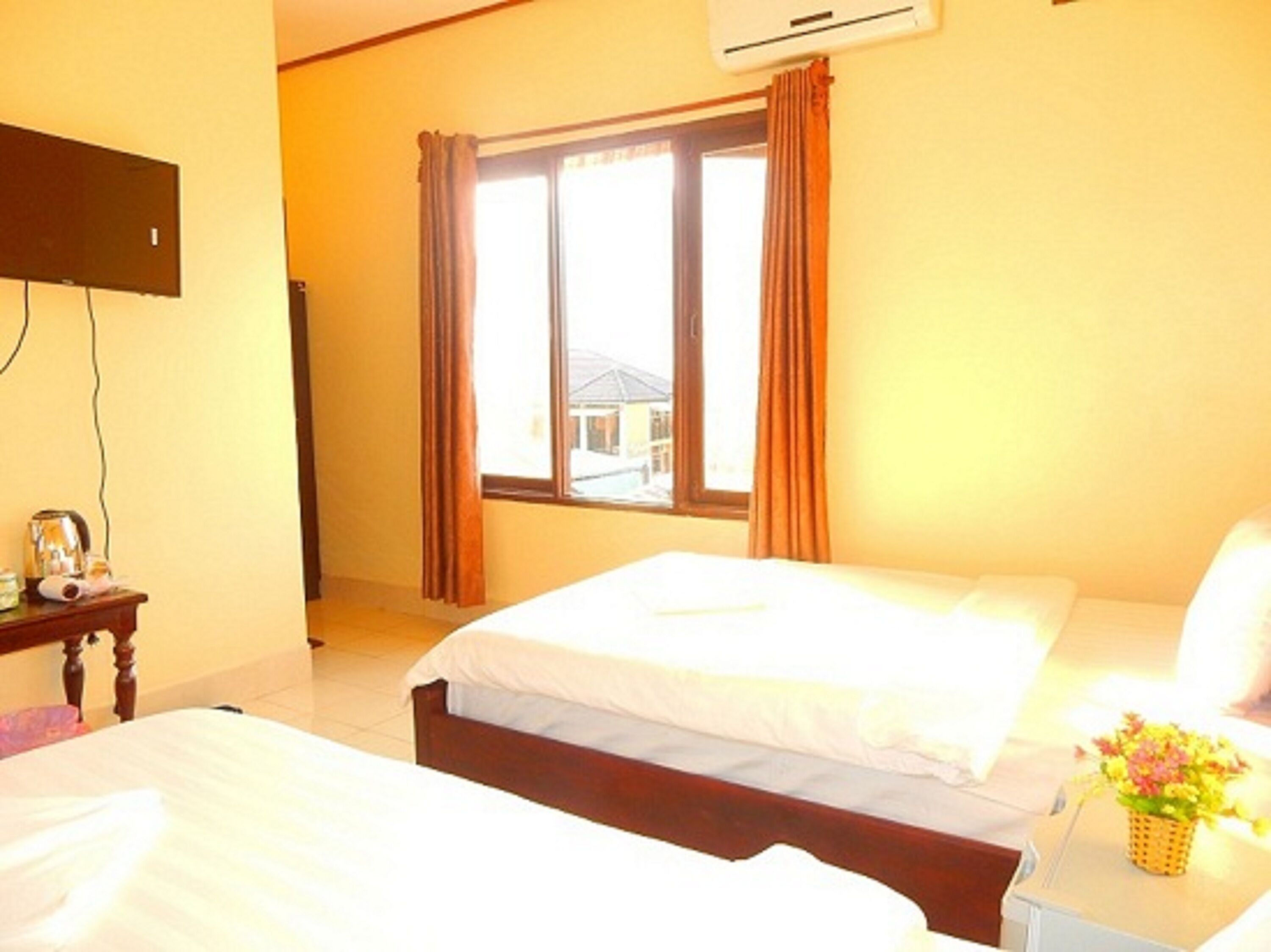A hotel room with two beds, a window with curtains, an air conditioning unit, and a small table with a kettle and a basket of flowers.
