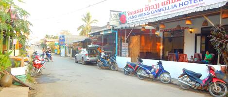 Vista de la calle con un hotel llamado Vang Vieng Camellia Hotel. Se ven motocicletas estacionadas al borde de la carretera, y algunas personas a lo lejos.