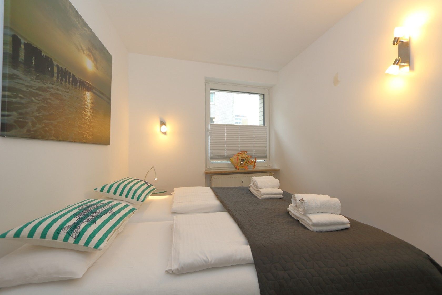 A hotel room with a bed, two pillows, a bedside table with a lamp, a window with blinds, and a wall-mounted light fixture.