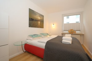 A bedroom with a bed, pillows, a bedside table, a window with blinds, a wall-mounted picture, and a small shelf with a fish decoration.