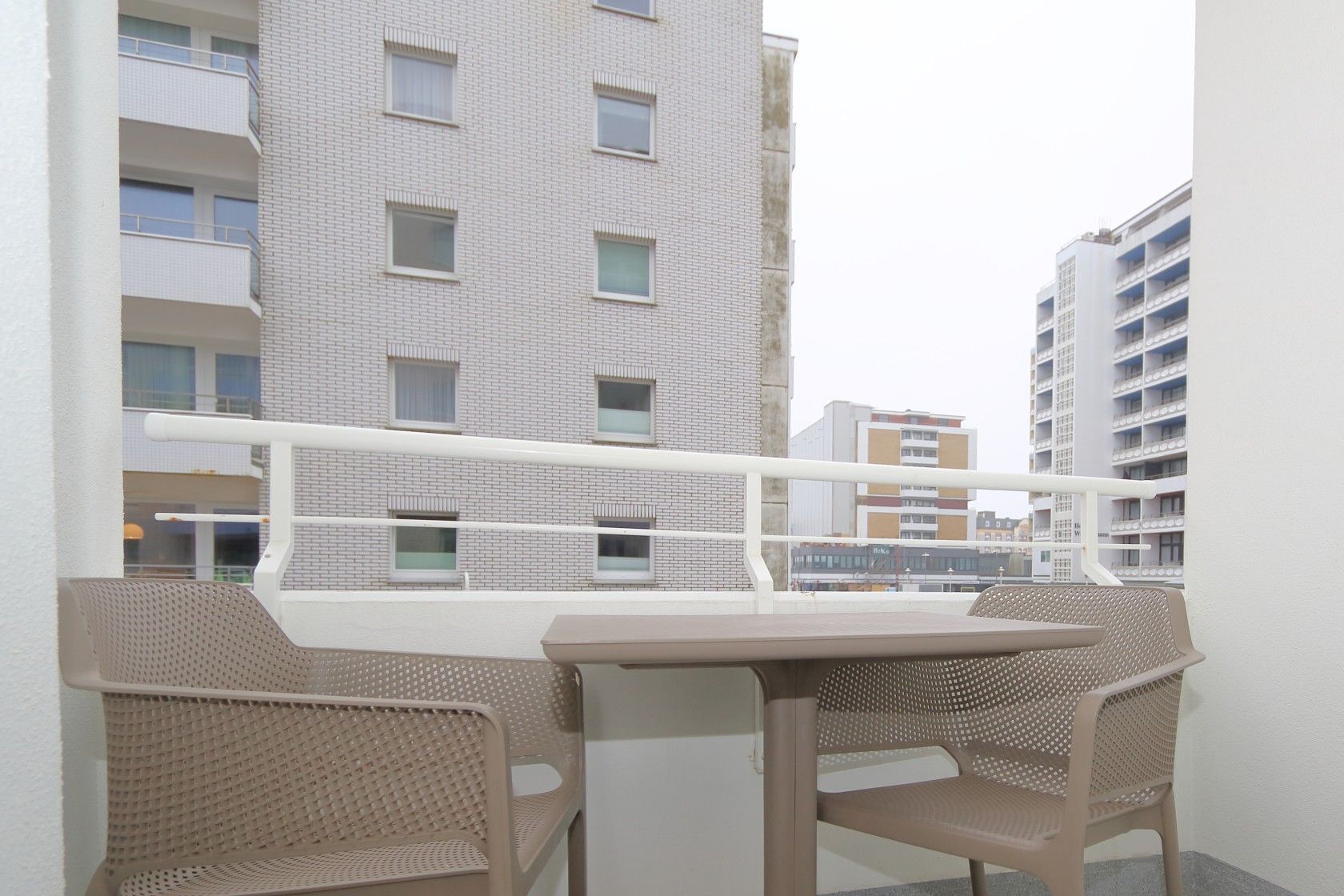Varanda com cadeiras de vime e uma mesa, com vista para um edifício com várias janelas e varandas.