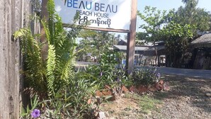 Une enseigne indiquant « BEAU BEAU BEACH HOUSE », avec une clôture en bois et des plantes au premier plan.