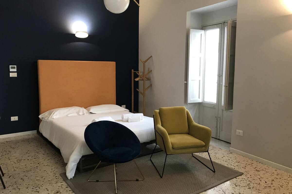 Una habitación de hotel moderna con una cama grande, un sillón amarillo, una silla azul oscuro, una lámpara de pie y una ventana con cortinas.