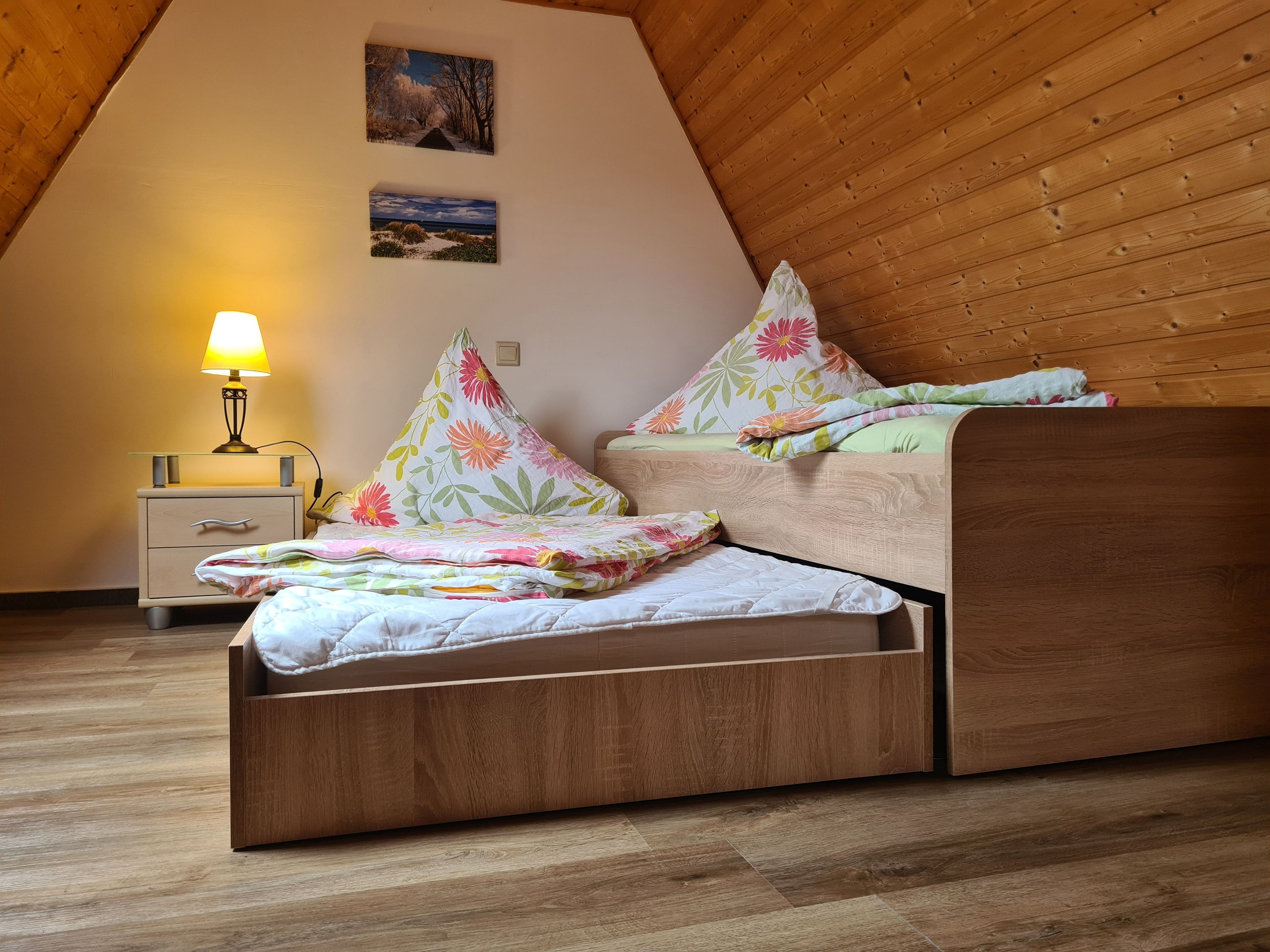 A loft-style bedroom with a wooden ceiling, a bed with floral bedding, a nightstand with a lamp, and two wall-mounted pictures.